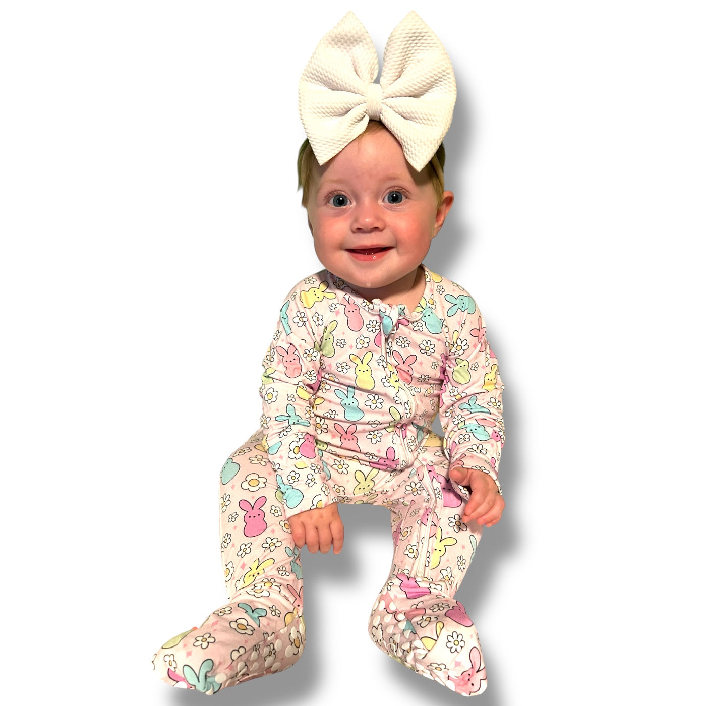 Baby wearing a colorful Easter Bunny zipper sleeper with a big white bow headband, smiling cheerfully.