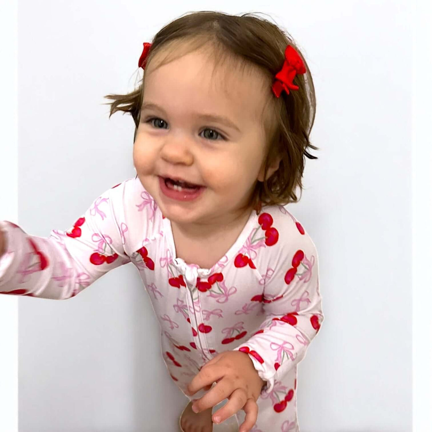Happy baby wearing Cherry Coquette bamboo zipper sleeper with cherry print, smiling and reaching out.