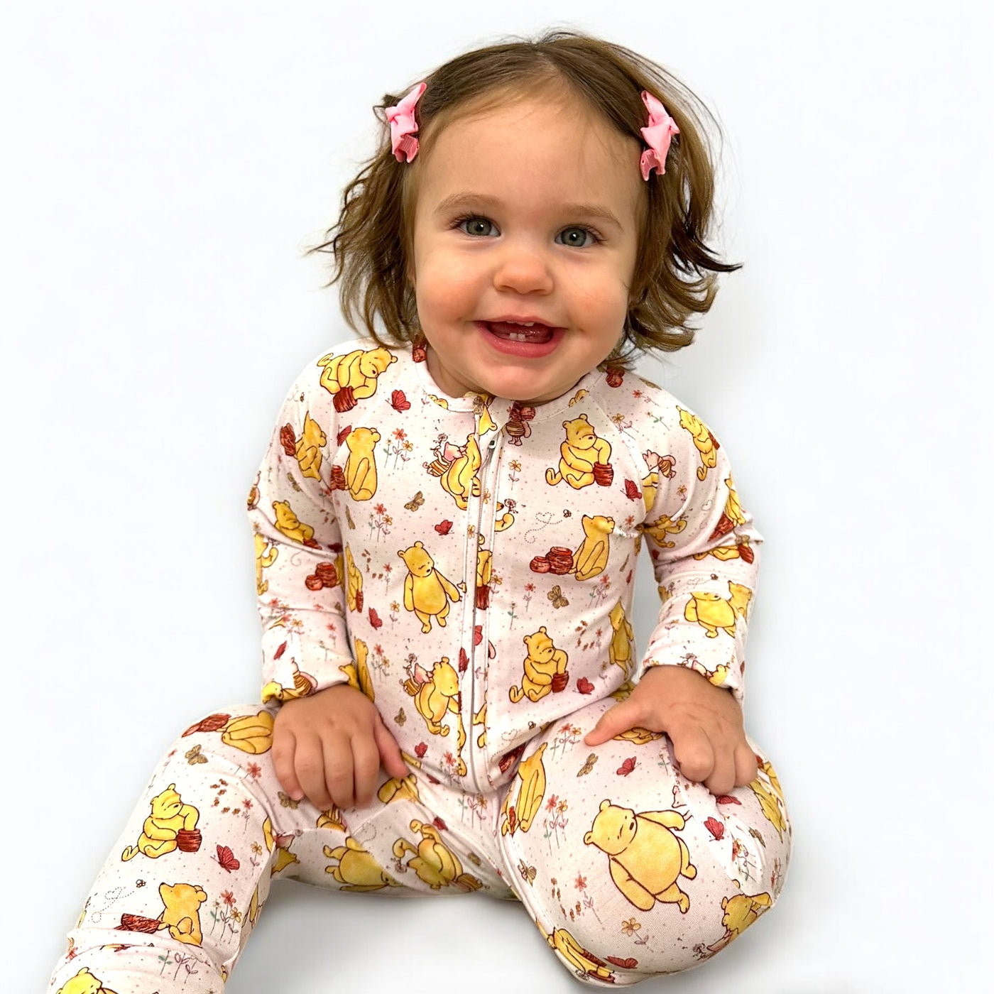 Toddler wearing a Classic Pooh zipper sleeper, smiling and sitting with bows in hair.