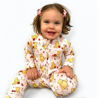 Toddler wearing a Classic Pooh zipper sleeper, smiling and sitting with bows in hair.