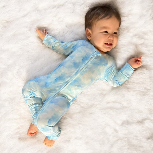 Baby wearing Blue Skies Bamboo Zipper Sleeper in a soft blue cloud print, lying on a fluffy white blanket.