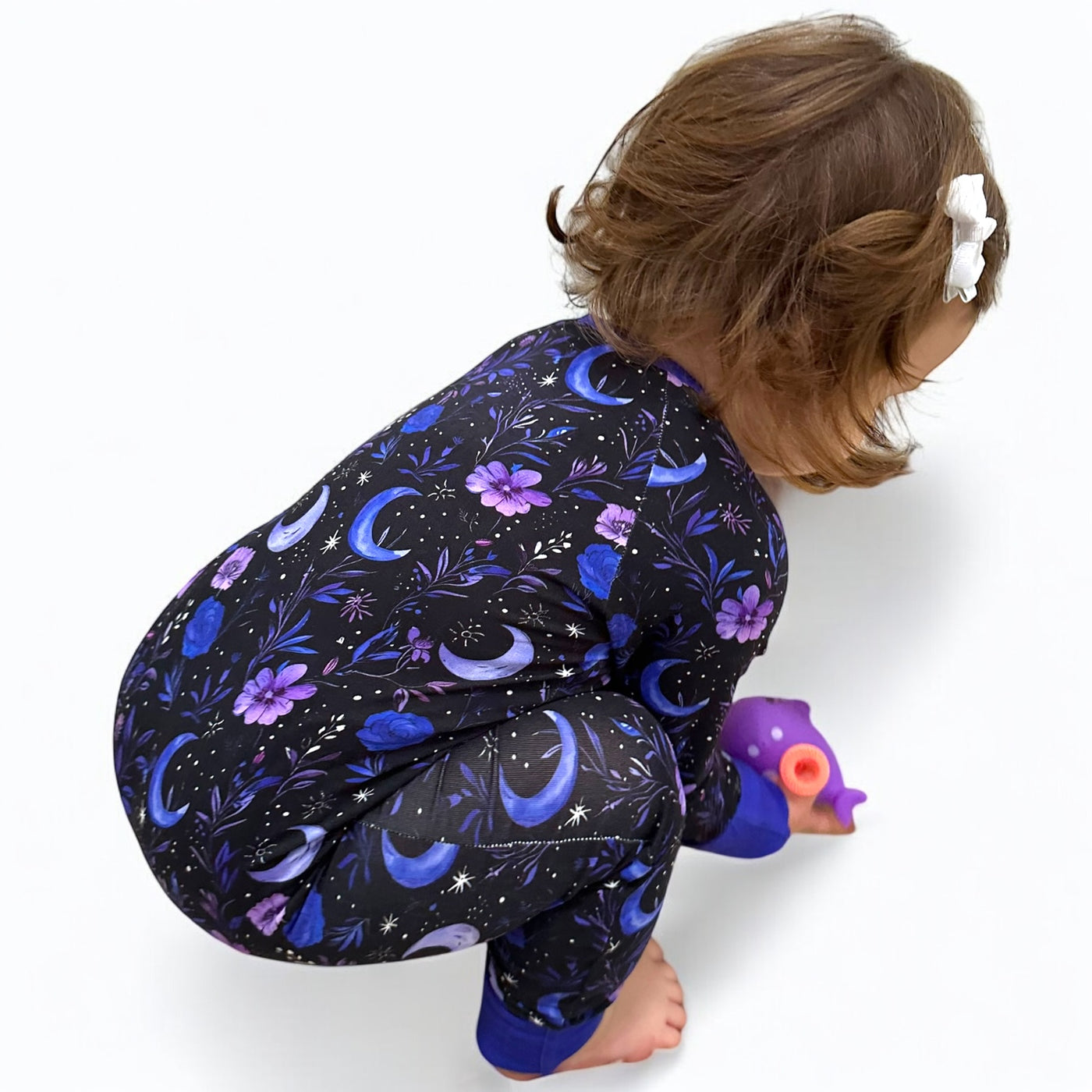 Toddler wearing a Purple Moonbeam bamboo zipper sleeper with floral and moon print, playing with a toy on the floor.