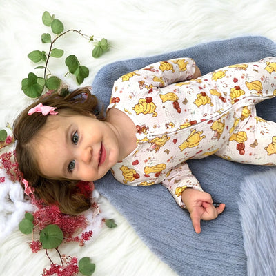 Smiling toddler in Classic Pooh zipper sleeper on a cozy blanket surrounded by soft greenery.