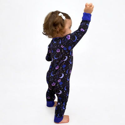 Baby wearing a Purple Moonbeam bamboo zipper sleeper with moons and stars design, raising one arm in playful joy.